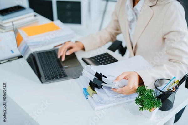 Obraz A young Asian businesswoman works alone at a desk with a laptop, stacks of papers and documents. She looks happy and successful managing accounts and preparing financial statements during the day.
