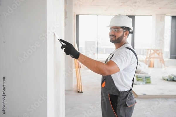 Fototapeta Indian Male builder in work overalls plastering a wall using a construction trowel.
