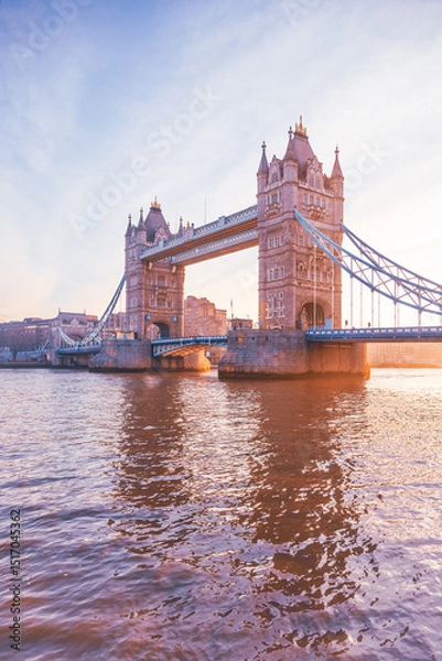 Fototapeta Tower Bridge stands majestically as the sun rises, casting warm hues over the Thames River. This iconic landmark is illuminated, showcasing its architectural beauty at dawn.