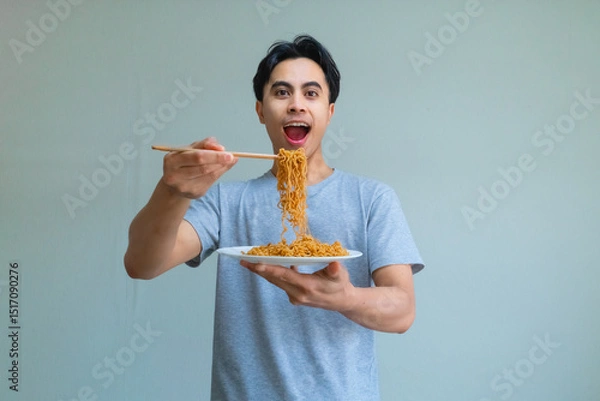 Fototapeta Happy young Asian man enjoying a plate of noodles with chopsticks, showing an excited expression on a clean background, perfect for food, culture, or lifestyle themes