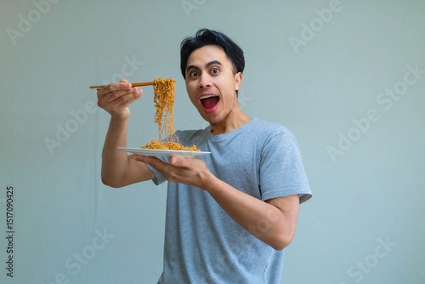 Fototapeta Happy young Asian man enjoying a plate of noodles with chopsticks, showing an excited expression on a clean background, perfect for food, culture, or lifestyle themes