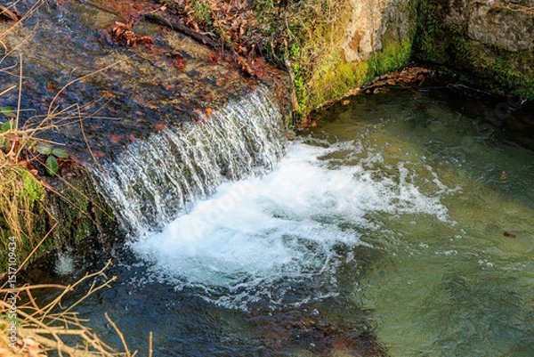 Fototapeta Serene small waterfall flowing into a clear stream through mossy rocks in nature
