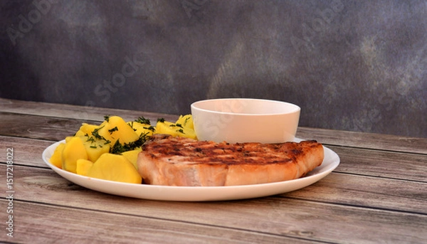 Obraz Screwed new potatoes and grilled pork entrecote on a round ceramic plate with a cup of spicy tomato sauce on a wooden table.