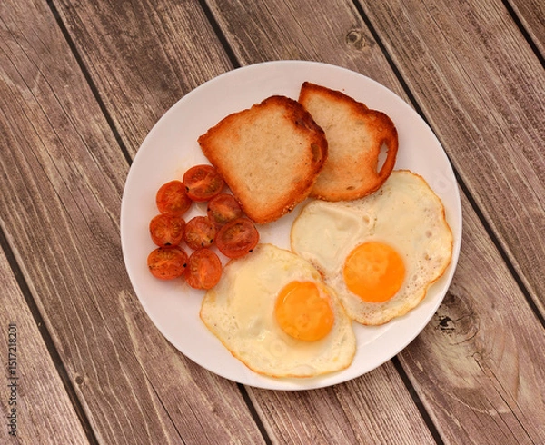 Obraz A plate of delicious breakfast, two fried eggs with cherry tomatoes and two wheat toasts on a wooden table.