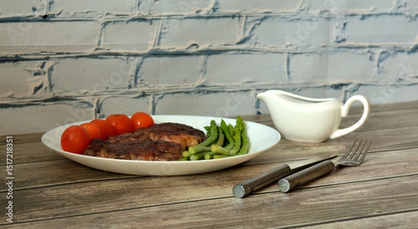 Obraz Two pieces of fried beef meat on a plate with cherry tomatoes and asparagus, sauce and cutlery on a light wooden table.