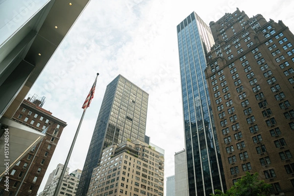 Obraz An American flag rises in front of towering buildings in Midtown Manhattan. Glass, brick, and stone architecture represents the city's diverse style.