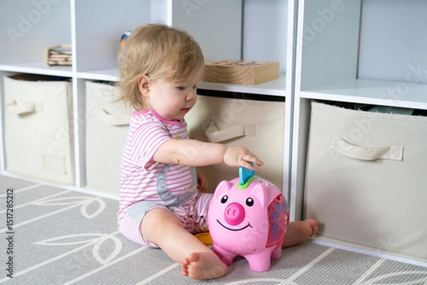 Fototapeta Cute baby girl sitting on nursery floor, playing with colorful piggy bank toy, early learning activities at home, toddler fun, indoor playtime, organized storage, happy childhood moments