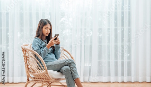 Fototapeta Young Asian Woman Relaxing in Modern Wicker Chair Near Window with Sheer Curtain, Happy Female Using Mobile Smartphone Enjoying at Home, Comfort and Cozy
