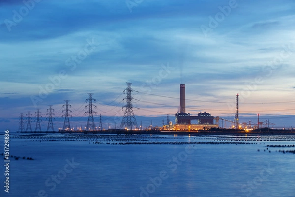 Obraz A coastal power plant is illuminated during twilight, with high-voltage transmission towers extending across the water. The scene captures industrial energy production and serene evening light.