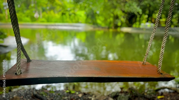 Fototapeta A close-up view of the swing shows its wooden seat smooth and quiet, hanging in front of a still pond bordered by the calm presence of trees.