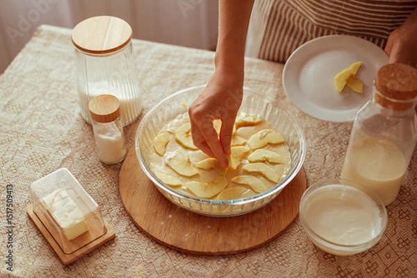 Fototapeta A cook in a striped apron arranges apple slices on top of batter in a glass pie dish, with baking ingredients on the table.