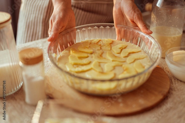 Fototapeta A cook in a striped apron holds a glass pie dish filled with apple slices arranged over batter, ready to be baked.