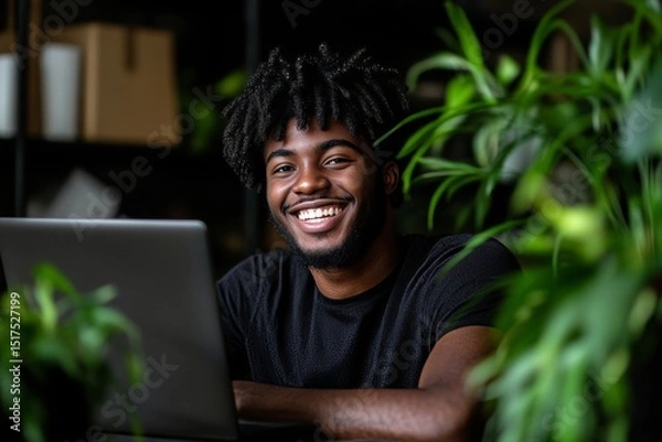 Fototapeta Young man smiling while working on laptop surrounded by indoor plants in a cozy home office setting