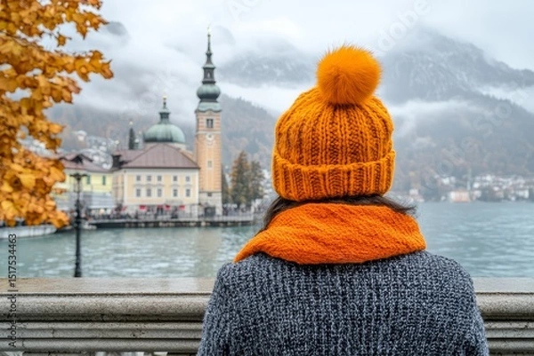 Fototapeta View of a woman wearing an orange hat and scarf enjoying the autumn scenery in a lakeside town with mountains in the background