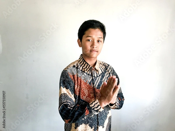 Obraz A teenage boy wearing a batik shirt doing a respectful greeting with hands together, isolated on white