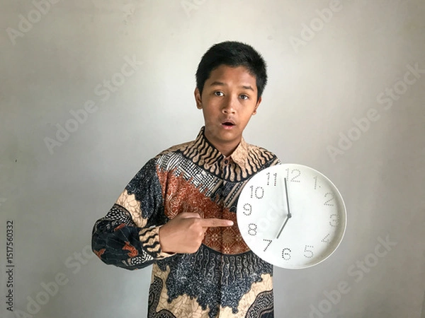Fototapeta A teenage boy wearing a batik shirt with a surprised expression holding a wall clock showing 7 o'clock, isolated on white