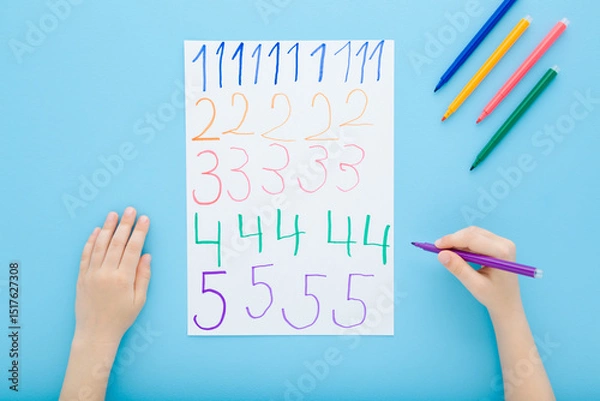 Fototapeta Little child boy hand holding color pen and writing colorful different numbers on white paper on blue table background. Pastel color. Doing school exercise. Closeup. Point of view shot. Top down view.