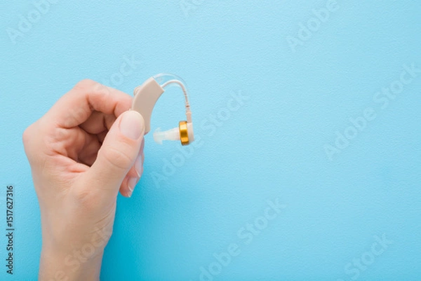 Fototapeta Young adult woman hand fingers holding and showing hearing aid on light blue table background. Pastel color. Closeup. Empty place for text. Top down view.