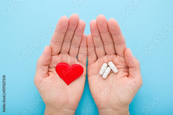 Fototapeta Young adult woman opened palms holding white capsule pills and red heart shape on light blue table background. Closeup. Pastel color. Receiving medicaments. Point of view shot. Top down view.