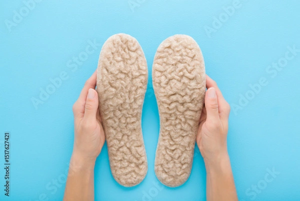 Fototapeta Young adult woman hands holding and showing new light soft natural sheep wool insoles on light blue table background. Pastel color. Closeup. Point of view shot. Top down view.