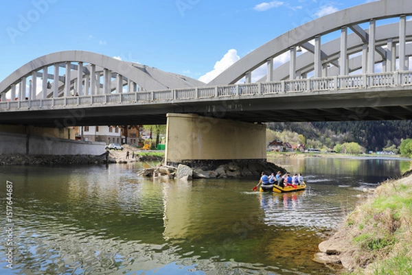 Fototapeta SZCZAWNICA, POLAND - APRIL 19, 2025: Tourists on a pontoon float down the river under the bridge.