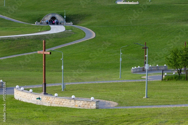 Fototapeta Multiple wooden crosses along a winding stone path in a manicured green field. Pilgrims approach an underground chapel in the background