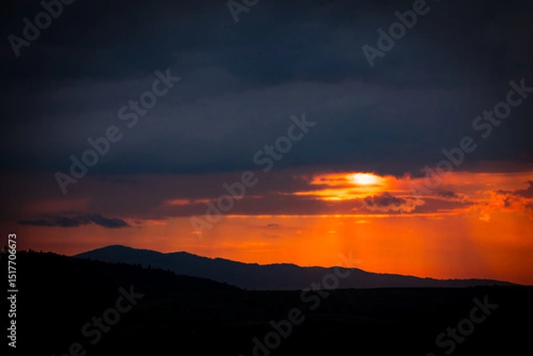 Fototapeta The last light of day filters through heavy clouds and showers, casting radiant streaks across a brooding sky above a layered mountainous backdrop — nature’s quiet yet powerful spectacle