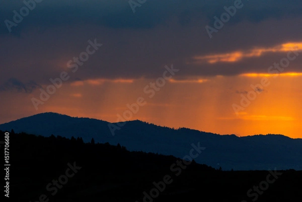 Fototapeta A fiery sunset breaks through dark clouds, painting the horizon in vibrant hues of orange and red while light rain gently falls over distant mountain ranges — a scene of serene wilderness