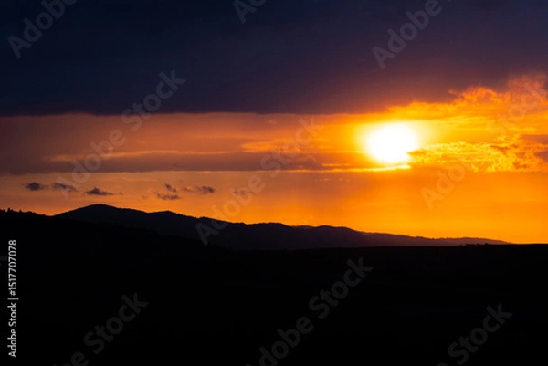 Fototapeta A radiant sunset over layered hills with a glowing orb descending behind cloud lines