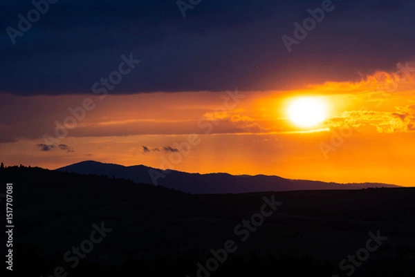 Fototapeta Intense sun framed by clouds as it sets beyond a forested mountain range.