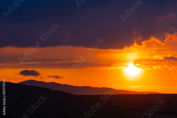 Fototapeta Evening sun blazes above forested ridges, illuminating thick clouds with fiery highlights and a deep amber sky