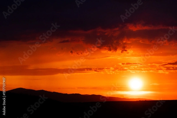 Fototapeta Bright golden sun hangs low over shadowed hills as vivid orange light paints the sky in waves of color and warmth