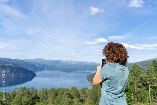 Fototapeta Young woman in her 30's seen from behind, capturing a stunning Norwegian of innvikfjorden fjord landscape with her mobile phone on a sunny summer day, enjoying the breathtaking scenic view