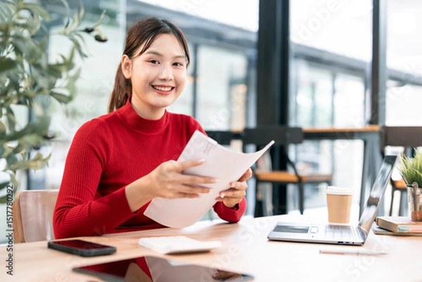 Obraz Shot of an attractive businesswoman in red sweater holding papaerwork sitting at her desk in an office.