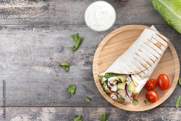 Fototapeta A delicious grilled chicken wrap with vegetables and avocado on wooden table. Top view. Copy space