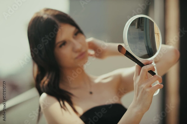 Obraz Young woman in a black strapless top applies makeup with a brush while looking in a small round mirror. The scene takes place during the day, with natural light illuminating the subject