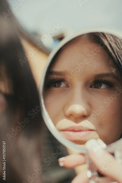 Obraz A young woman checks her appearance using a mirror, seemingly outdoors. The light emphasizes her features and the scene gives off a modern and fashionable vibe