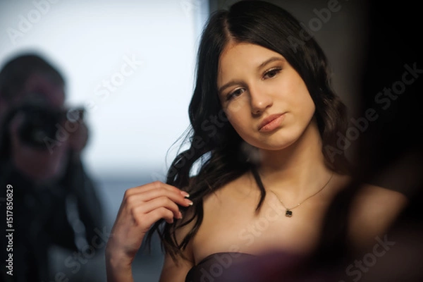 Obraz In a studio setting, a fashionable social media influencer with dark hair poses while a photographer takes pictures. She wears a strapless top and a necklace