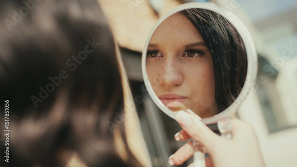 Obraz A young woman looks into a handheld mirror while applying makeup. She is fashionable and carefully poses for an upcoming social media post