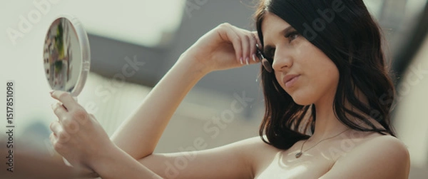 Obraz A young woman looks at herself in a hand mirror. She poses, perhaps for content on social media, in a modern, urban environment. She seems fashionable and modern