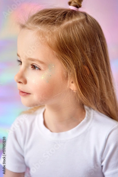 Fototapeta A close-up portrait of a charming little girl with long blond hair. She looks thoughtfully into the distance. The model's face is decorated with glitter. The background has gentle pastel shades.