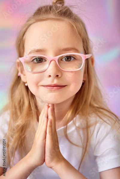 Fototapeta A girl with long hair and pink glasses. She put her hands together in a prayer pose. The girl's smiling, wearing a white T-shirt. The background is soft pastel shades, creating a pleasant, dreamy mood