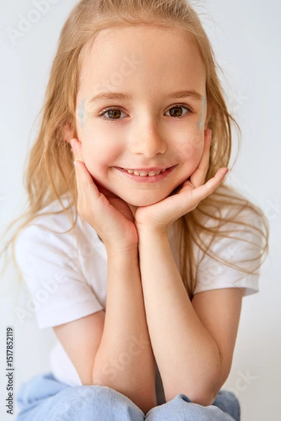 Fototapeta A charming young girl with long blond hair smiles radiantly, resting her head on her hands. She is dressed in a white T-shirt and light trousers. There are delicate blue patterns on her cheeks