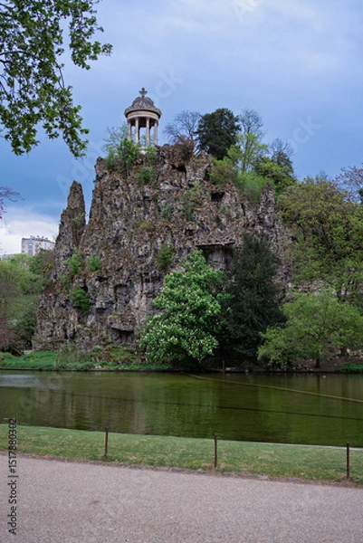 Fototapeta Temple de la Sibylle on a rocky cliff in Buttes-Chaumont Park, Paris. Scenic view with trees, lake, and dramatic sky. Iconic romantic landscape in spring