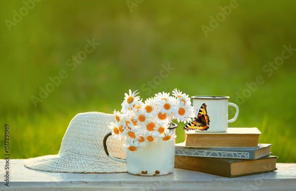 Fototapeta summer nature background. chamomile flowers in mug, butterfly, books and braided hat close up on table in garden. Beautiful rustic floral composition. relaxation, harmony atmosphere. soft focus