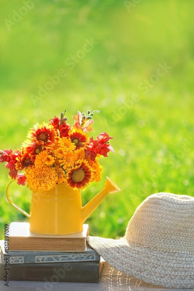 Fototapeta bright flowers bouquet in yellow watering can and books on table in garden, natural background. romantic atmosphere nature image. relax time. composition with flowers. summer season.