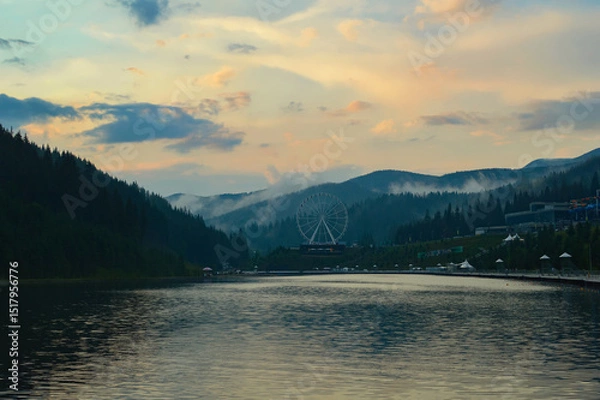 Obraz A picturesque view of the lake and mountains during sunset, Bukovel ski resort in the Carpathians. Fog drifts between the mountain peaks after rain. Ferris wheel in the background.