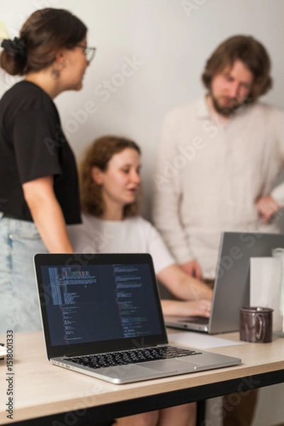 Fototapeta Three software developers brainstorming in a modern office. Team coding session with laptops showing code, planning web development and design. Tech startup environment and teamwork
