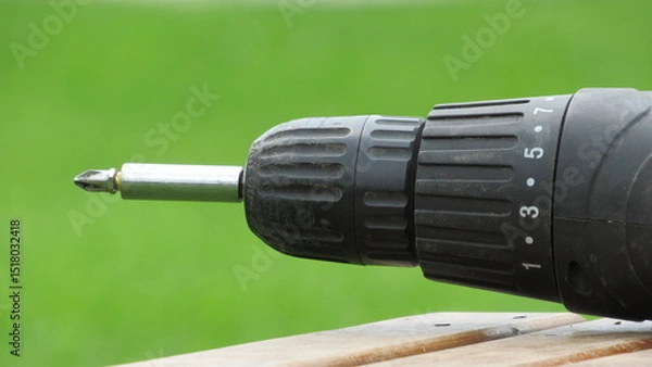Obraz Close-up view of a black electric drill resting on a wooden surface in a green outdoor setting during daylight