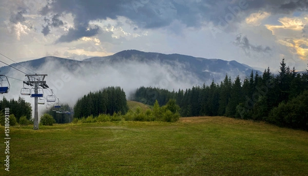 Obraz Cableway in the Carpathian mountains. The sun comes out from behind the storm clouds after the rain. A thick fog rolls over the forest and mountains.